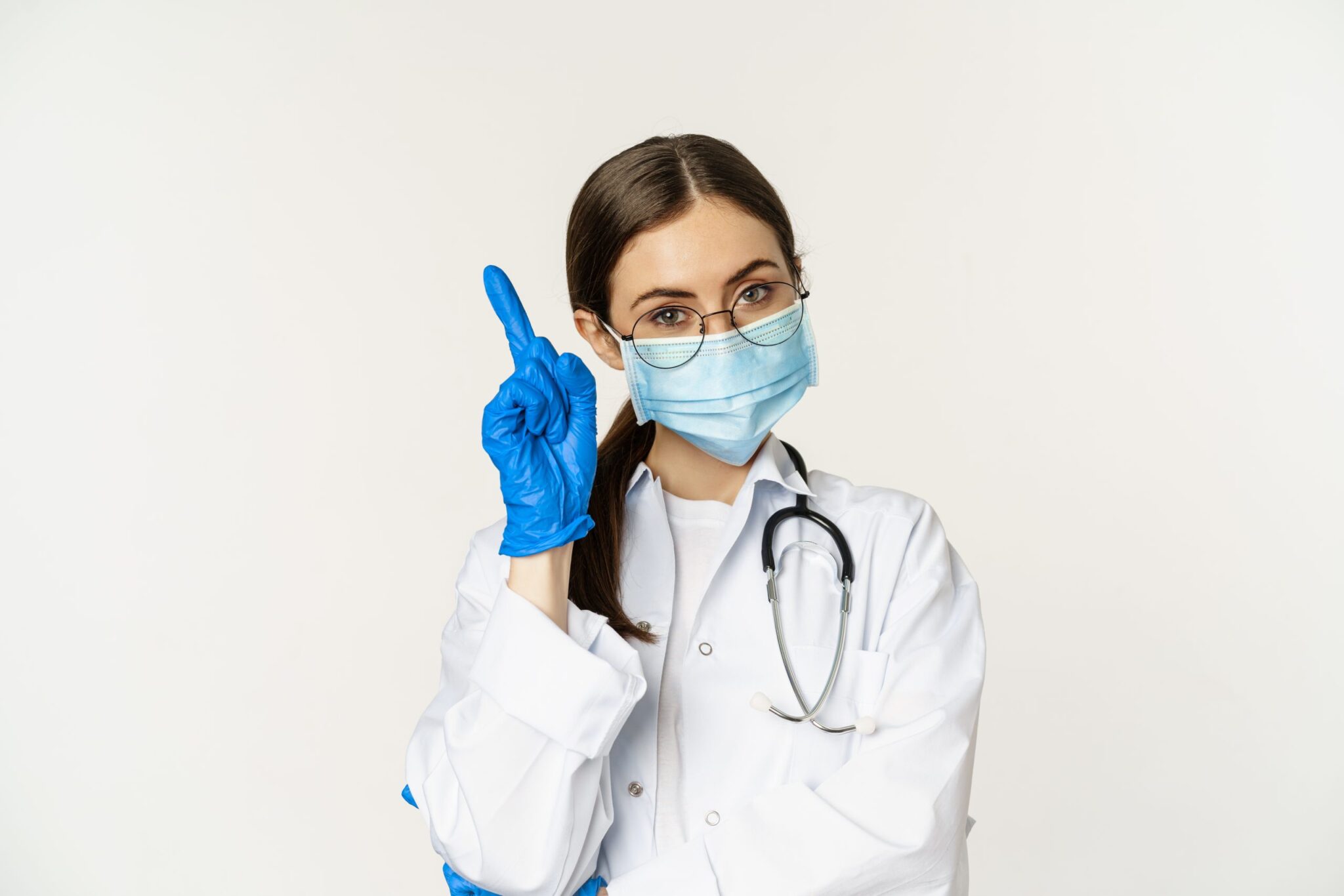 Close up portrait of female doctor in glasses, medical face mask and gloves, pointing finger up at link, clinic logo, advertisement on top, standing over white background